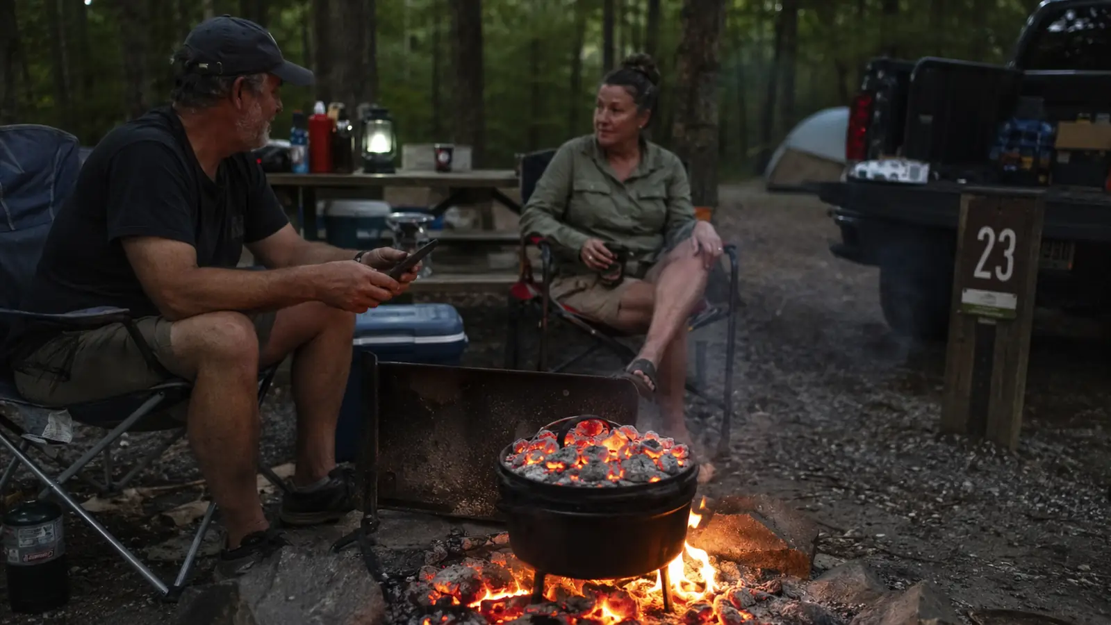A Lodge cast iron camp dutch oven sitting in a bed of glowing campfire coals with more coals piled on the flanged lid