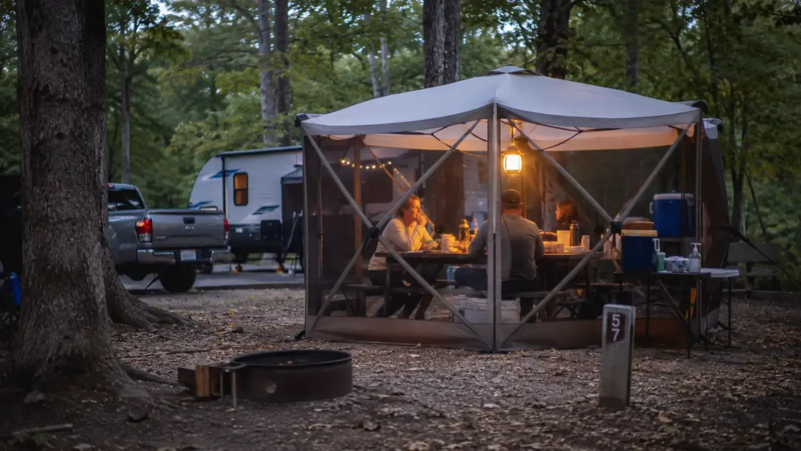 Alvantor pop up screen house tent set up at a wooded campsite at dusk with mesh walls glowing from a lantern inside